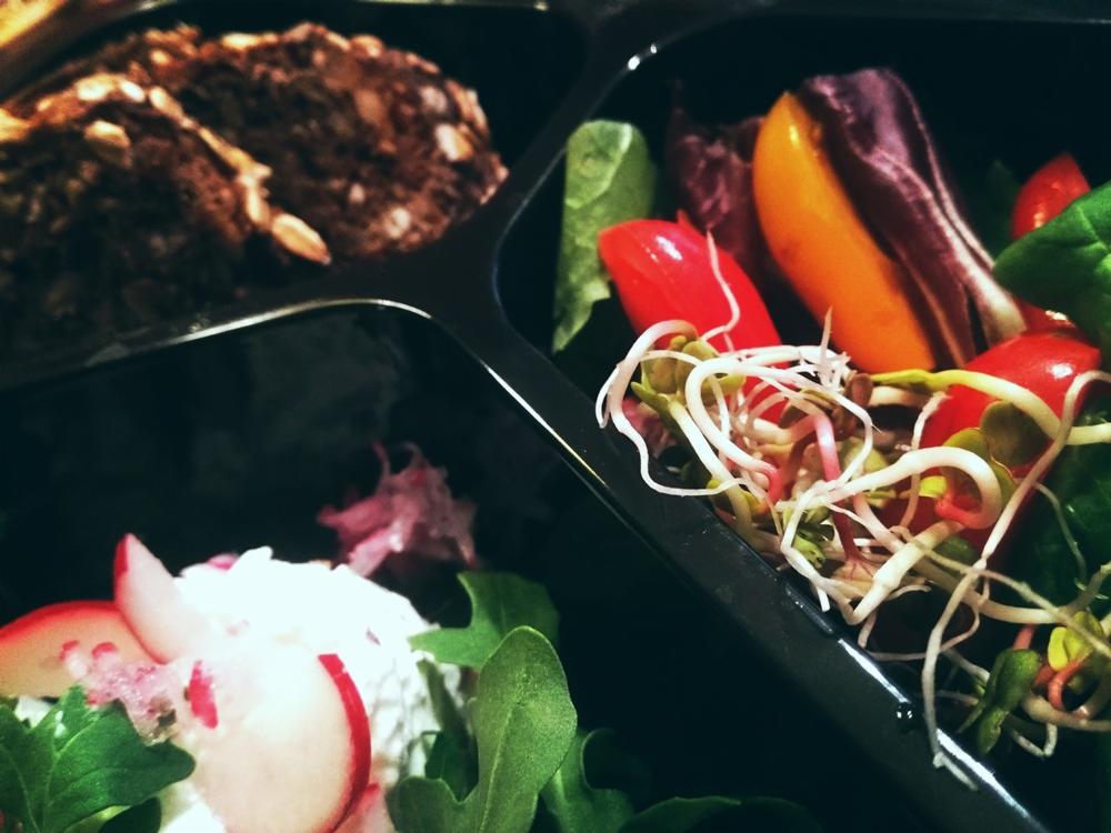 Czterodzielna czarna tacka z różnorodnymi, zdrowymi przekąskami: ciemny chleb z ziarnami, sałatka z rzodkiewką i serem, mieszanka sałat z papryką, pomidorami i kiełkami.