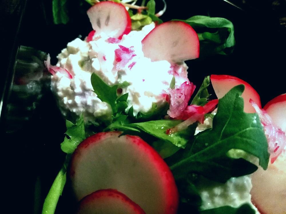 Sałatka z białym serem, rzodkiewką i zielonymi listkami, podana na czarnym talerzu, z bliska, w ciemnym oświetleniu.