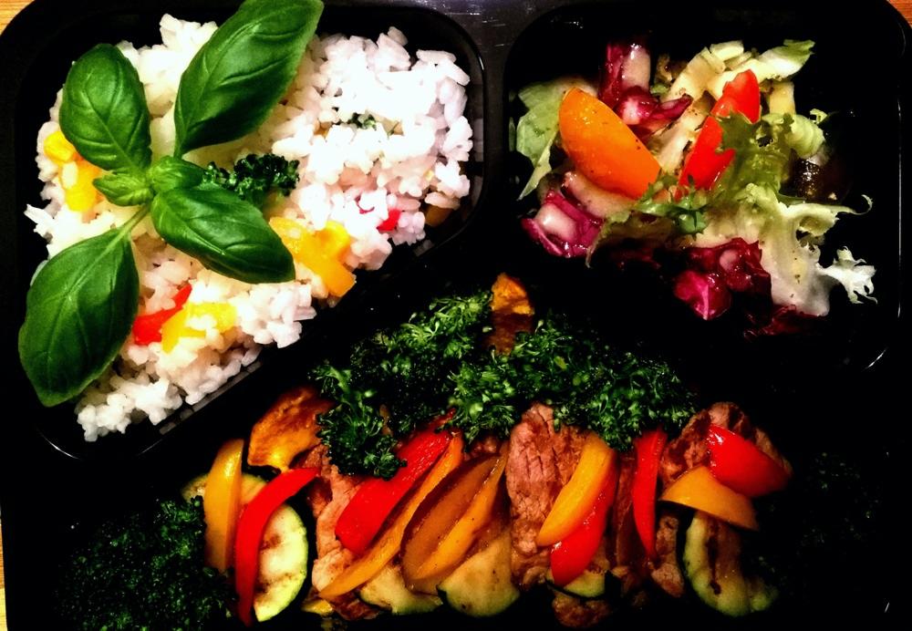 Czarny pojemnik z trzema przegródkami zawierającymi: ryż z warzywami i bazylią, sałatkę mieszaną z pomarańczą i mięso z grillowanymi warzywami, posypane zieloną pietruszką.