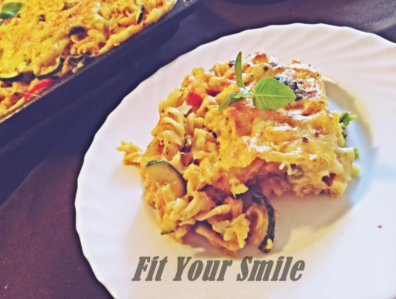 Porcja zapiekanki makaronowej z warzywami i serem na białym talerzu, udekorowana listkiem bazylii, obok blaszka z resztą zapiekanki. Widoczny napis 'Fit Your Smile'.