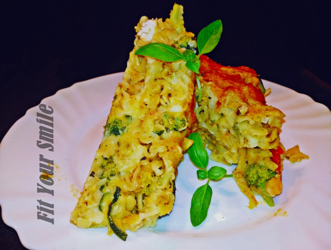 Dwa trójkątne kawałki zapiekanki makaronowej z brokułami i cukinią, udekorowane świeżymi listkami bazylii, na białym talerzu z napisem 'Fit Your Smile', na ciemnym tle.