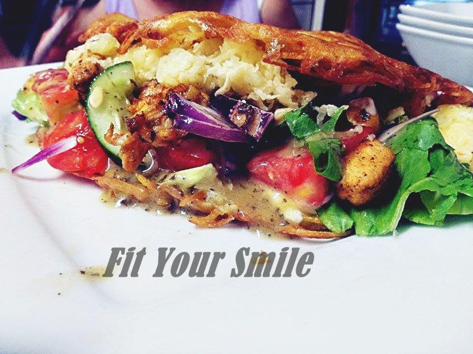 Otwarta kanapka z warzywami, w tym pomidorami, ogórkiem, czerwoną cebulą i sałatą, polana sosem, z widoczną chrupiącą warstwą wierzchnią i napisem 'Fit Your Smile'.