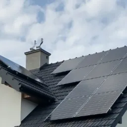 Panele słoneczne na dachu domu jednorodzinnego z ciemną dach&oacute;wką i jasną elewacją, komin z dwoma zakończeniami w kształcie ptak&oacute;w, błękitne niebo z chmurami w tle.