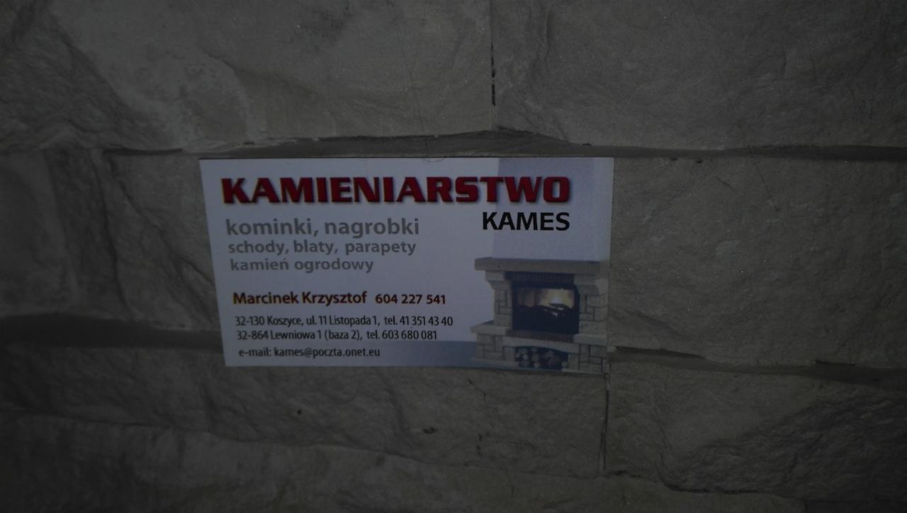 Zdjęcie wizytówki firmy kamieniarskiej 'KAMES' na tle ściany imitującej kamień, oferującej kominki, schody, blaty, parapety i inne wyroby z kamienia.