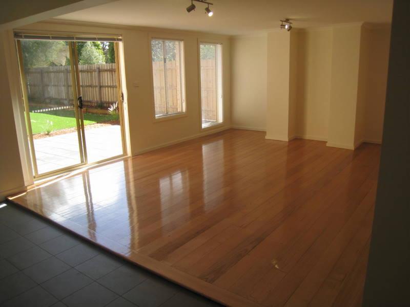 Puste pomieszczenie z parkietem, widoczne okno przesuwne z wyjściem na zewnątrz, gdzie znajduje się trawnik i ogrodzenie. Wnętrze jasne, z trzema oknami z żaluzjami. Podłoga przechodzi...