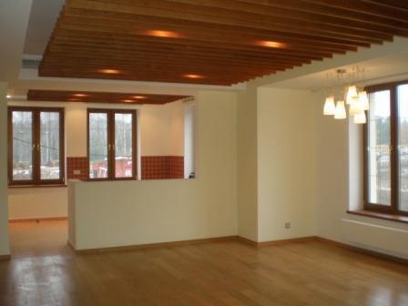 Jasne wnętrze z drewnianą podłogą i ażurowym podwieszanym sufitem z oświetleniem punktowym. W tle widoczna kuchnia z oknami i czerwonymi płytkami na ścianie.