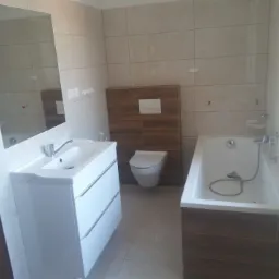 Wykończona łazienka z beżowymi płytkami, białą umywalką z szafką, wanną obudowaną drewnopodobnymi panelami i toaletą w zabudowie, widok zza drzwi.