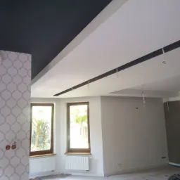 Wnętrze w trakcie wykończenia: pomalowany na czarno sufit sąsiaduje z białym, podwieszanym sufitem z zamontowaną szyną oświetleniową; ściany częściowo pomalowane na szaro, tapeta w geometryczny...