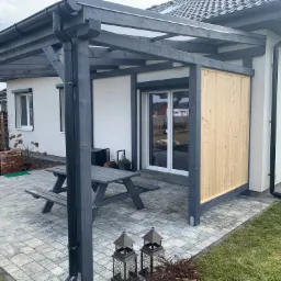 Zadaszona przestrzeń wypoczynkowa z drewnianą ławką i stołem na wyłożonym kostką brukową patio przylegającym do białego domu z szarą stolarką, ozdobne lampiony na pierwszym planie.