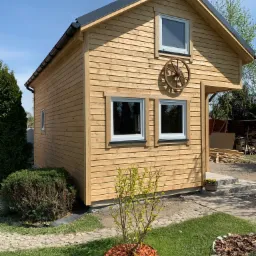 Niewielki drewniany domek letniskowy z jasnych desek, z dwuspadowym dachem i dekoracyjnym kołem od wozu na elewacji, otoczony zielonym trawnikiem z elementami dekoracyjnymi z kory.