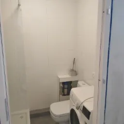 Wykończona łazienka z kabiną prysznicową, toaletą w zabudowie i pralką, wyłożona białymi, kwadratowymi płytkami.