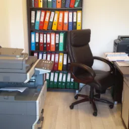 Biuro z dużym regałem wypełnionym kolorowymi segregatorami, biurkiem z komputerem, drukarką i fotelem biurowym, obok stoi duża kopiarka z dokumentami.