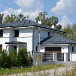 Dwukondygnacyjny dom z szarym dachem blaszanym, białymi ścianami i brązowymi roletami zewnętrznymi, ogrodzony czarnym płotem.