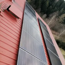 Panele słoneczne zamontowane na czerwonym, blaszanym dachu domu, z widocznym kominem i lasem w tle.