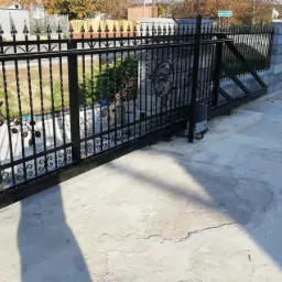 Automatyczna, przesuwna brama wjazdowa z czarnego metalu, zdobiona motywami roślinnymi, zamontowana na betonowym podłożu i słupkach z szarej kostki.