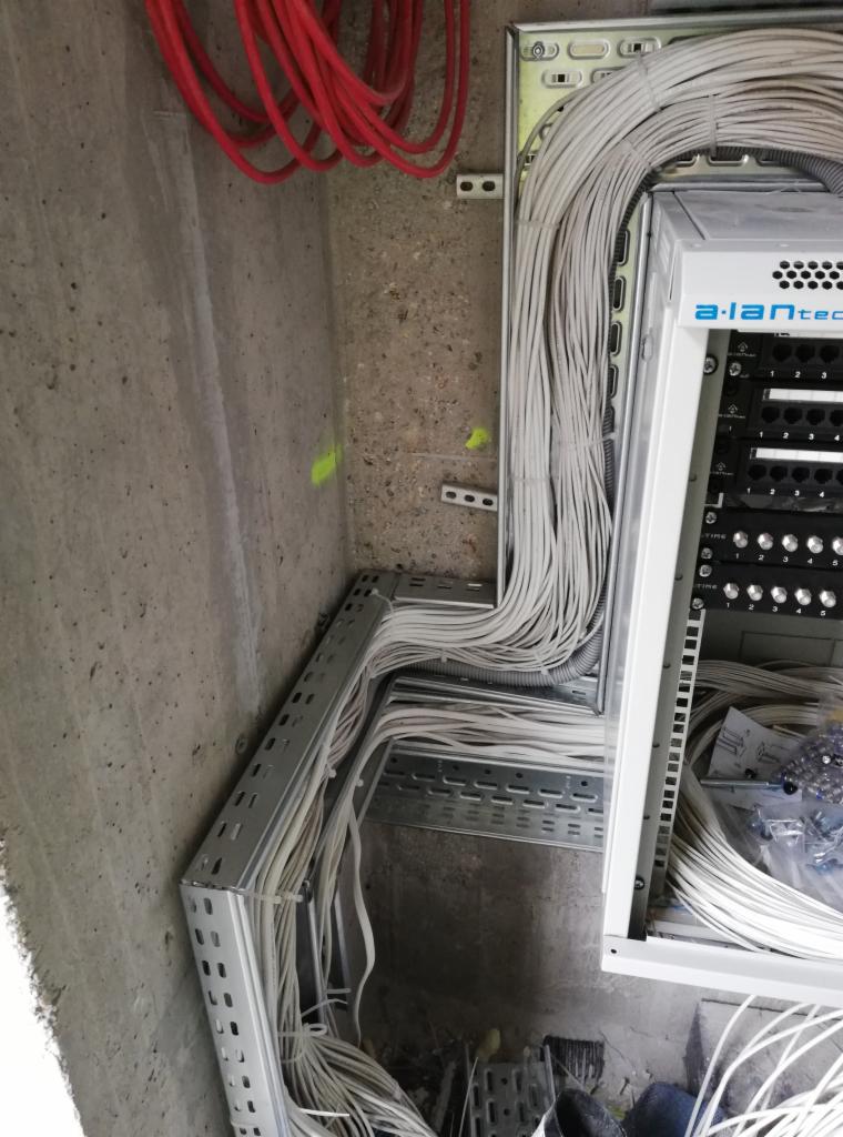 Instalacja kabli telekomunikacyjnych w korytach i szafie rack, widoczne kable białe i czerwone oraz elementy mocujące na surowym betonowym tle.