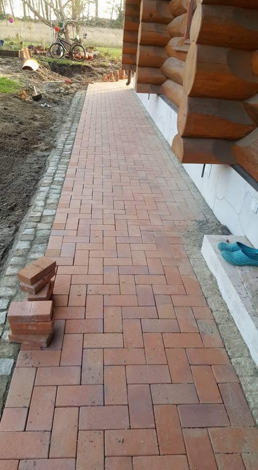 Czerwona kostka brukowa ułożona w jodełkę wzdłuż drewnianego domu, z widocznymi zapasowymi kostkami i przygotowanym podłożem.