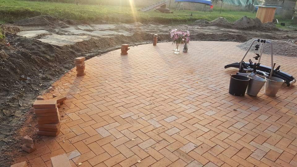 Półokrągły taras z ceglanej kostki ułożonej w jodełkę, w tle przygotowany teren pod dalsze prace ziemne, na gotowej powierzchni stoi wazon z kwiatami i akcesoria rowerowe.