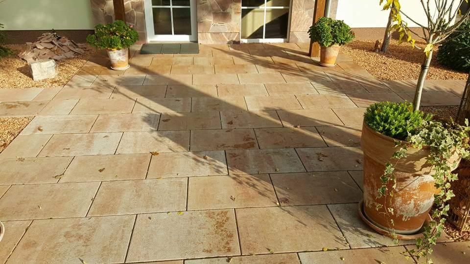 Ułożony, prostokątny chodnik z beżowo-brązowej kostki brukowej prowadzący do drzwi wejściowych, z donicami z roślinami po bokach i fragmentami ogrodu z kamykami.