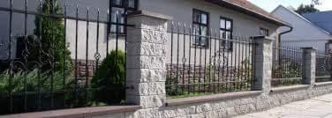 Metalowe ogrodzenie posesji z dekoracyjnymi elementami, osadzone na niskim murku z kamienną fakturą, z widocznymi słupkami z bloczków imitujących kamień.