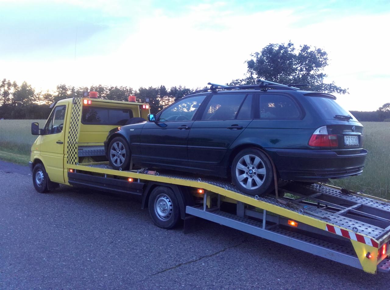 Żółta laweta z zielonym kombi BMW na platformie, widok z boku na tle pola i zachmurzonego nieba.