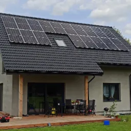 Dom jednorodzinny z zamontowanymi panelami fotowoltaicznymi na czarnym dachu, widoczne okno dachowe, drewniany taras z meblami ogrodowymi i skrzynka z narzędziami.