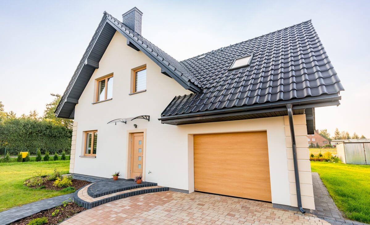 Nowoczesny dom jednorodzinny z garażem, jasną elewacją, drewnianymi oknami i drzwiami, oraz ciemną dachówką, widziany z poziomu podjazdu wyłożonego kostką brukową.