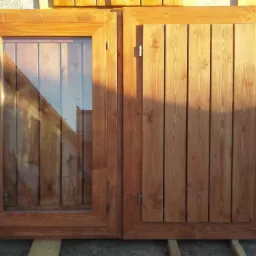 Dwa skrzydła okienne z drewna w odcieniu miodowym, jedno z szybą, drugie z drewnianymi panelami, przygotowane do montażu w Tomaszowie Mazowieckim.