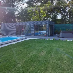 Nowoczesny budynek z szarej blachy falistej z przeszklonymi drzwiami przesuwnymi, przylegający do basenu i jacuzzi na zadbanym trawniku z białym żwirem dekoracyjnym i zielenią