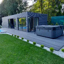 Nowoczesny, szary budynek modułowy z dużymi oknami, tarasem z desek kompozytowych i jacuzzi, otoczony zielonym trawnikiem i drzewami w słoneczny dzień.