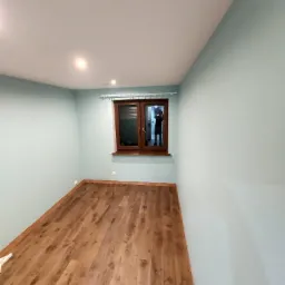 Pomalowane na pastelowo-niebiesko pomieszczenie z drewnianą podłogą z paneli, brązowym oknem i odbiciem osoby robiącej zdjęcie.