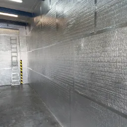 Wnętrze hali przemysłowej z zamontowanymi płytami warstwowymi o srebrnej, metalicznej powierzchni, widoczna drabina aluminiowa z czarno-żółtymi oznaczeniami.