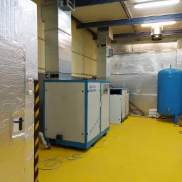 Wnętrze hali produkcyjnej z urządzeniami CompAir DEMAG, instalacją wentylacyjną i zbiornikiem ciśnieniowym na żółtej posadzce.