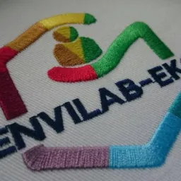 Zbliżenie na kolorowe logo 'ENVILAB-EKO' wyhaftowane na białym materiale, z widoczną strukturą splotu tkaniny i detalami ściegów.