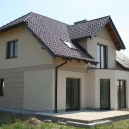 Dwukondygnacyjny dom jednorodzinny z brązową dachówką, jasną elewacją z poziomym pasem dekoracyjnym, widoczne okna dachowe i taras z kostki brukowej na trawniku.