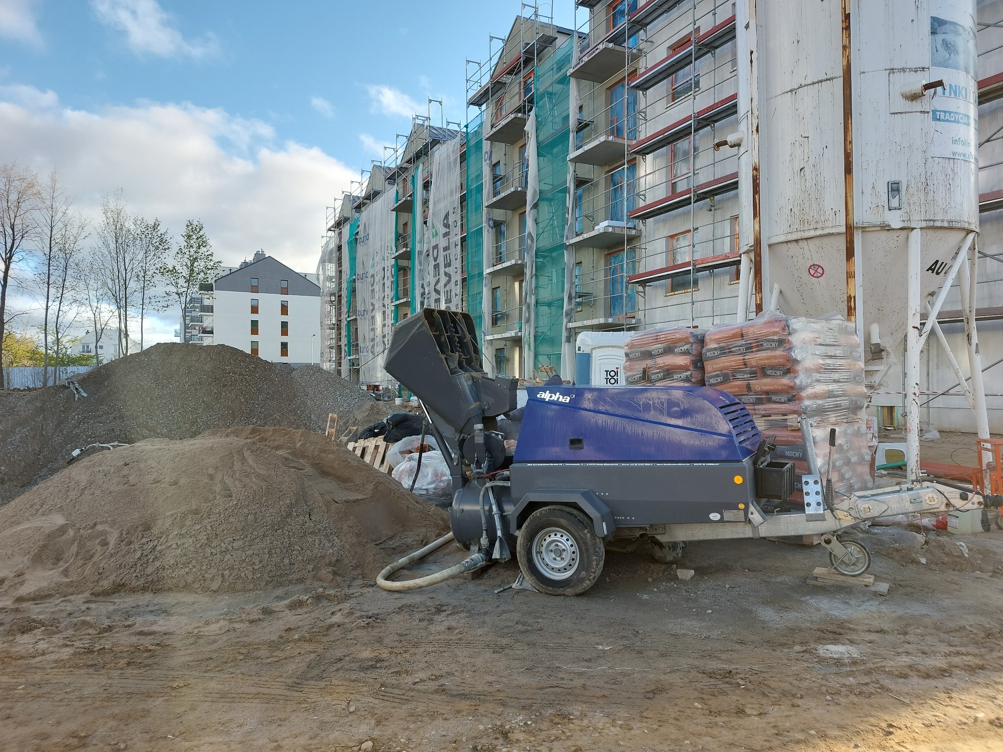 Agregat do wylewek Alpha na budowie wielorodzinnej, obok hałda piasku i worki z cementem. W tle budynek w rusztowaniach i biały budynek mieszkalny.