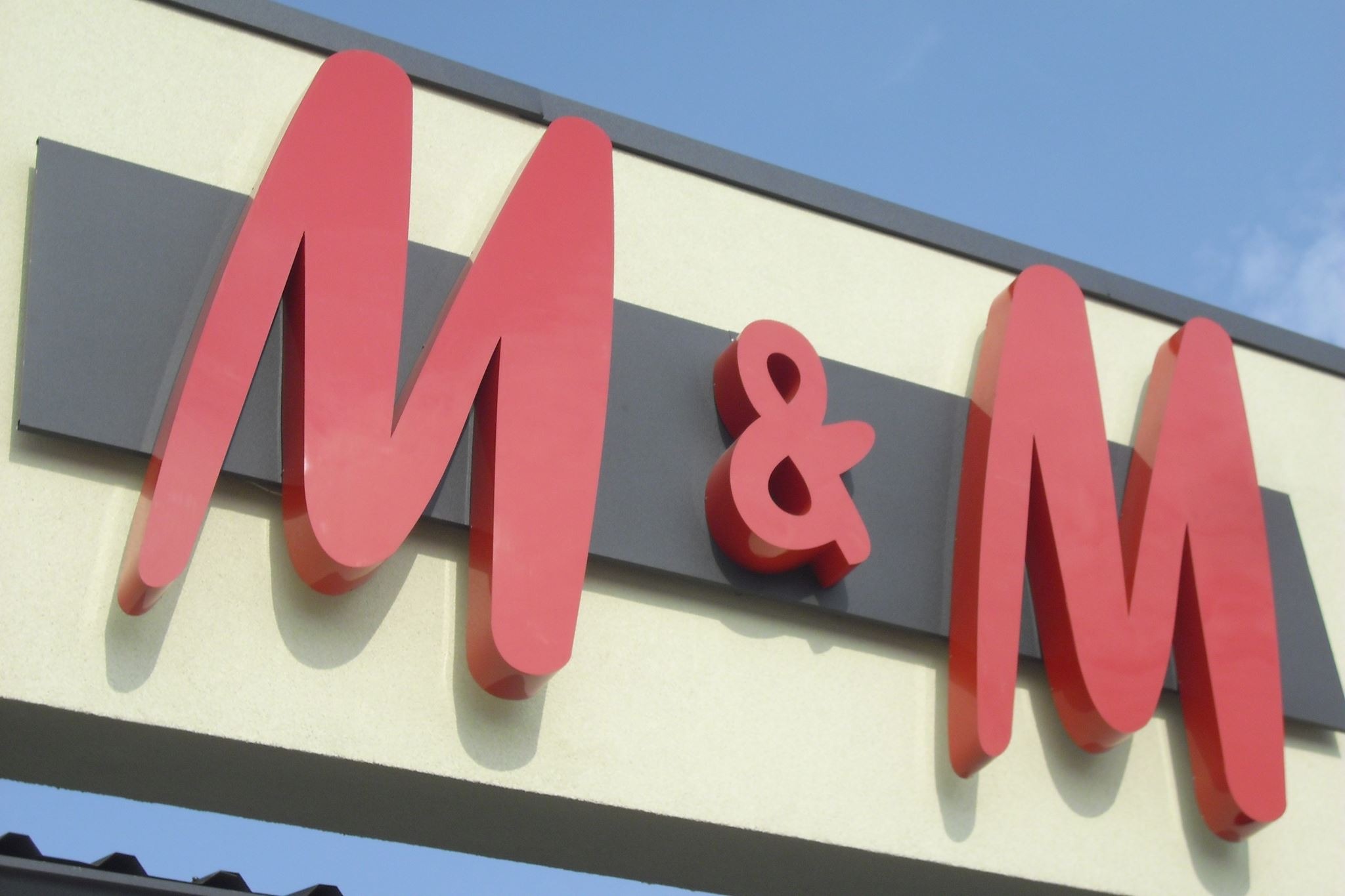 Czerwone, przestrzenne litery M&M zamontowane na szarym panelu na elewacji budynku, widoczne z dołu pod kątem, z fragmentem dachu w dolnej części kadru.