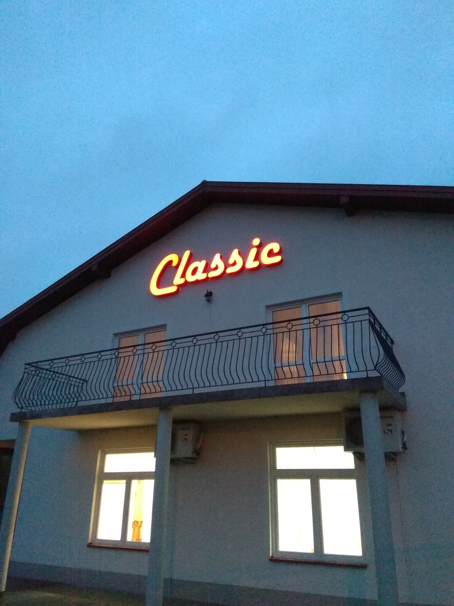 Elewacja budynku z balkonem i neonowym napisem 'Classic' świecącym ciepłym światłem na tle wieczornego nieba.