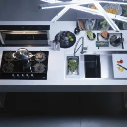 Nowoczesna kuchnia z białym blatem, czarną płytą grzewczą z garnkiem, zlewem ze świeżymi ziołami, deską do krojenia z warzywami i designerskim oświetleniem sufitowym.