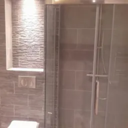 Szara łazienka z kabiną prysznicową ze szklanymi przesuwanymi drzwiami, podświetloną wnęką z dekoracyjną okładziną ścienną i podwieszaną toaletą z chromowanym przyciskiem spłukującym.