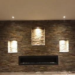 Ściana z kamienia dekoracyjnego z trzema wnękami oświetlonymi punktowo i czarnym wkładem kominkowym, widoczne płytki dystansowe na podłodze.