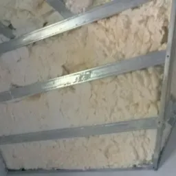 Ocieplanie poddasza pianką PUR między metalowymi profilami konstrukcyjnymi, widoczna struktura pianki i szara ściana boczna.