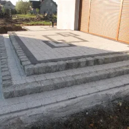 Schody i chodnik wyłożone szarą kostką granitową z ciemnoszarym obramowaniem i geometrycznym wzorem przed budynkiem z roletami.