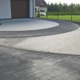 Zakrzywiony podjazd z jasnoszarej kostki brukowej obramowany ciemniejszą kostką, prowadzący do garażu i ogrodzenia z zielonym trawnikiem w tle.