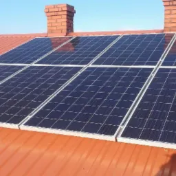 Panele słoneczne na czerwonym dachu domu z ceglanym kominem w tle, widok z góry.