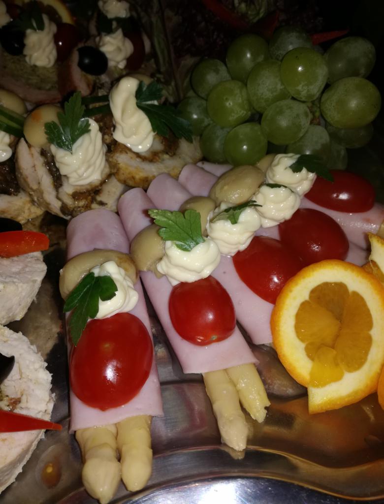 Dekoracyjna taca z przekąskami: szparagi owinięte szynką z pomidorkami koktajlowymi i kleksem majonezu, roladki mięsne z kremem, zielone winogrona i plasterki pomarańczy.