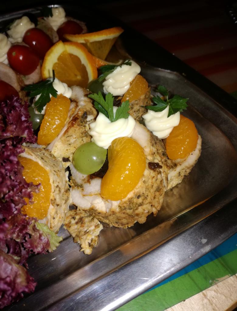 Rolada z kurczaka dekorowana mandarynką, winogronami, sosem majonezowym i natką pietruszki, podana na srebrnej tacy obok pomidorków koktajlowych i sałaty.