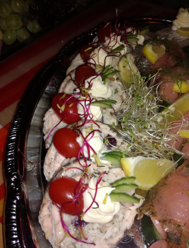 Elegancka srebrna patera z roladkami rybnymi udekorowanymi pomidorkami koktajlowymi, kleksem majonezu, ogórkiem, kiełkami i cytryną, część zastawy stołowej widoczna w tle.