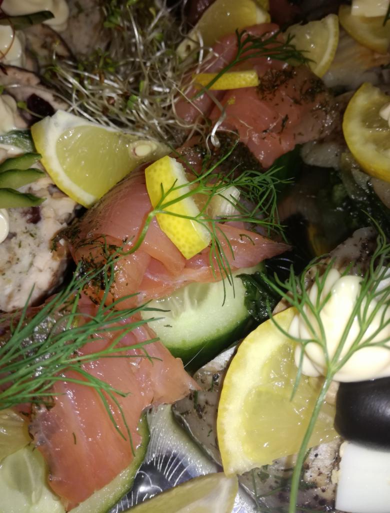 Zbliżenie na talerz z wędzonego łososia, plasterków cytryny, świeżego ogórka, zieleniny i innych przekąsek, całość udekorowana koperkiem.