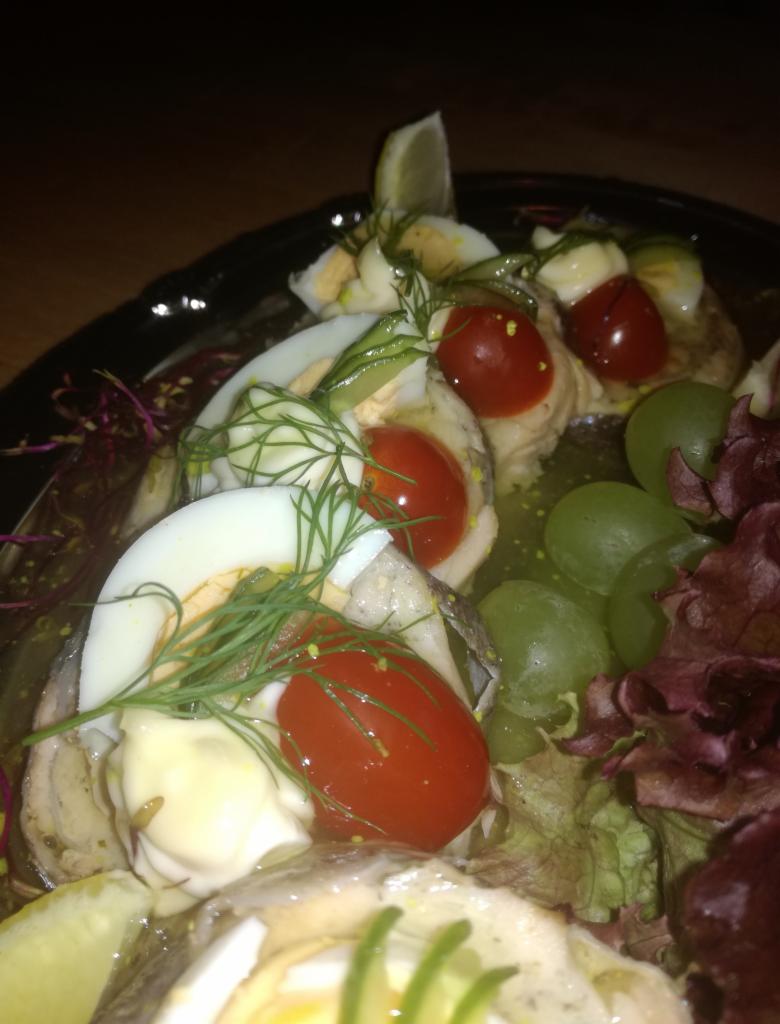 Śledź w oleju, udekorowany jajkiem na twardo, pomidorkami koktajlowymi, koperkiem, winogronami i sałatą na czarnym talerzu.